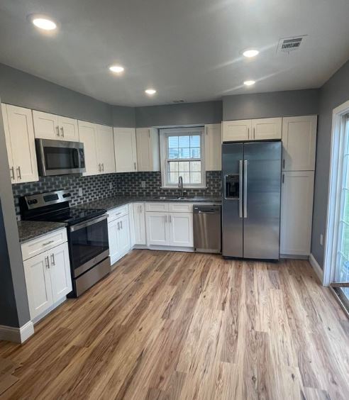 An open concept kitchen without island, L-Shape layout, bright, white, shaker cabinets, stainless steel appliances, wood floor. 